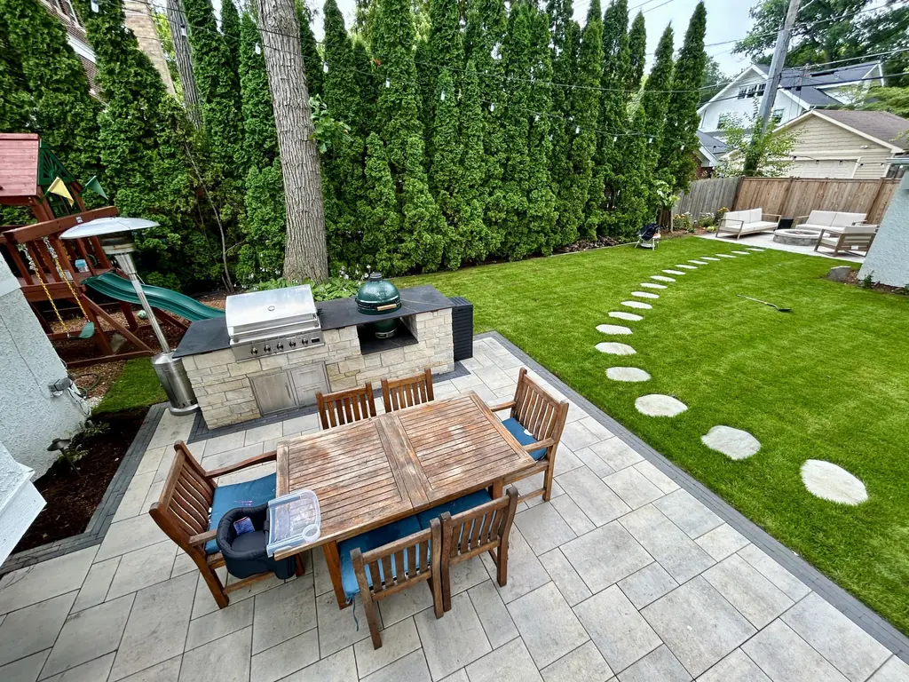 Outdoor kitchen and dining table on a paver patio beside a lawn